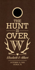 A cornhole board with a circular cutout at the top, featuring the phrase 'THE HUNT IS OVER' in beige lettering, antler graphics, the names 'Elizabeth & Albert,' and the date/location 'OCTOBER 12, 2030 / Ocala, FL.'.