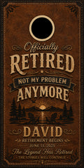 A square brown leather-look retirement sign or print with decorative typography reading 'Officially RETIRED NOT MY PROBLEM ANYMORE' and 'DAVID', plus a black circular cutout near the top and a scenic forest sunset illustration near the bottom.
