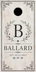 Personalized cornhole board with a distressed white wood background, featuring a black circular hole near the top, a large letter B enclosed in a leaf wreath, the name BALLARD, and the date EST. 05.