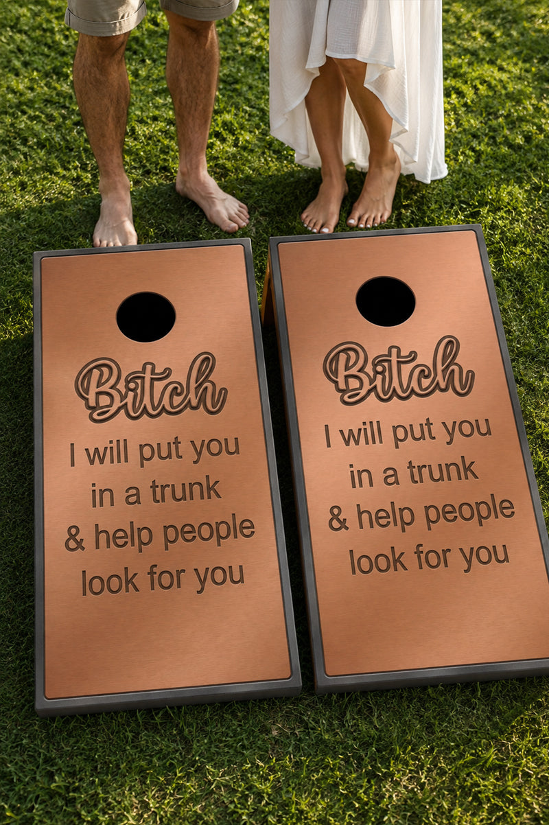 Two rectangular cornhole boards with dark gray frames and black holes, printed with the phrase 'Bitch I will put you in a trunk & help people look for you' in dark lettering, placed on green grass with bare feet and a white dress visible above them.