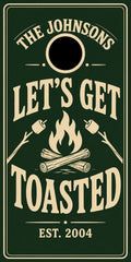 A square dark green sign or print with cream text reading “THE JOHNSONS LET’S GET TOASTED EST. 2004,” plus a cream campfire graphic with marshmallows and logs.