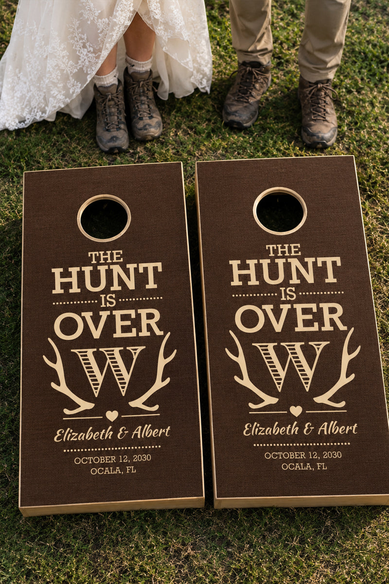 Personalized cornhole boards on a grassy lawn with a bride's dress and a man's hiking boots visible at the top, featuring the text 'THE HUNT IS OVER' with antlers and the names 'Elizabeth & Albert' and the date 'October 12, 2030'.