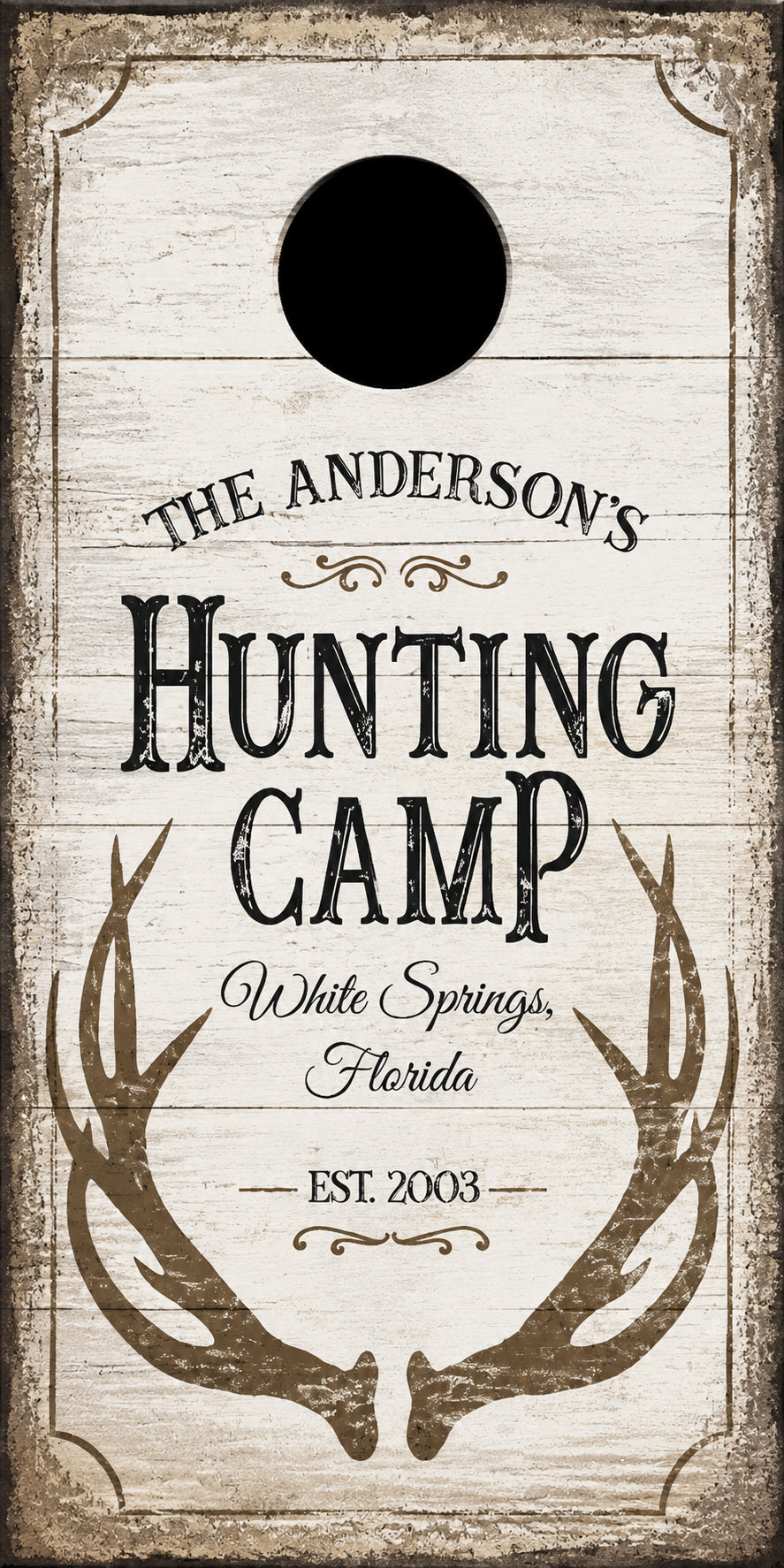 A personalized cornhole board with a rustic white wood-grain finish and distressed brown border. The board features a circular hole near the top, with the text 'THE ANDERSON'S HUNTING CAMP White Springs, Florida EST.