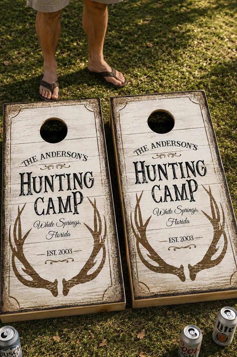 Two personalized cornhole boards on a grassy lawn, with a person's legs and flip-flops visible above them. The boards are printed with rustic whitewashed wood texture, antler graphics, and the text 'The Anderson's Hunting Camp White Springs, Florida' and 'Est.
