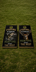 Two cornhole boards with hunting and fishing-themed graphics placed on a green grass lawn. The boards feature black backgrounds with fish and deer illustrations, gold text reading 'FISHING MY PROBLEMS' and 'HUNTING SOLVES THE REST,' plus a circular gold emblem near the top of each board.