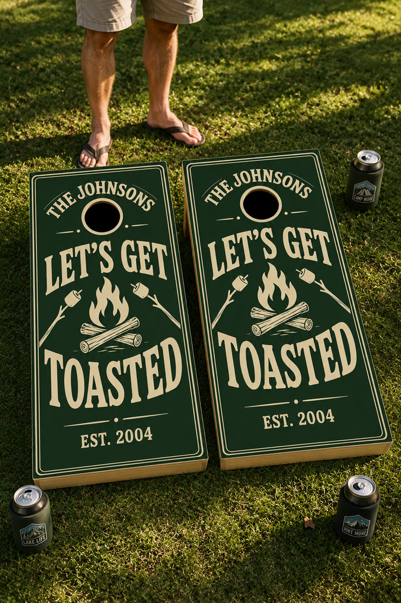 Two personalized cornhole boards on a grassy lawn, with a person's legs and feet visible at the top. The boards are dark green with cream lettering that reads 'THE JOHNSONS LET'S GET TOASTED EST.