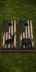 Two rustic cornhole boards placed on green grass. Each board has a circular hole and features a distressed American flag design with a black deer silhouette on one board and a black bear silhouette on the other.