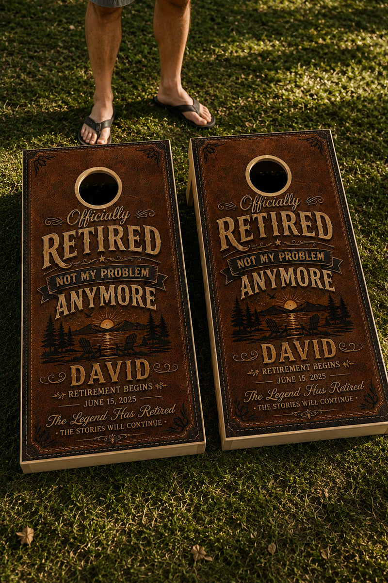 Two personalized cornhole boards on a grassy lawn, with a person's legs and feet visible standing behind them. The boards have a distressed brown wood-grain finish and printed text reading 'Officially Retired Not My Problem Anywhere' with a lake and trees illustration and the name 'David'.