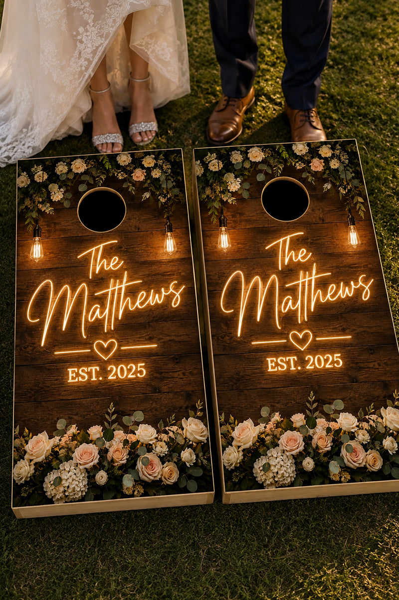 Two personalized cornhole boards on a grassy lawn at night, decorated with floral designs and warm glowing string lights. The boards feature the custom text 'The Matthews' and 'EST.
